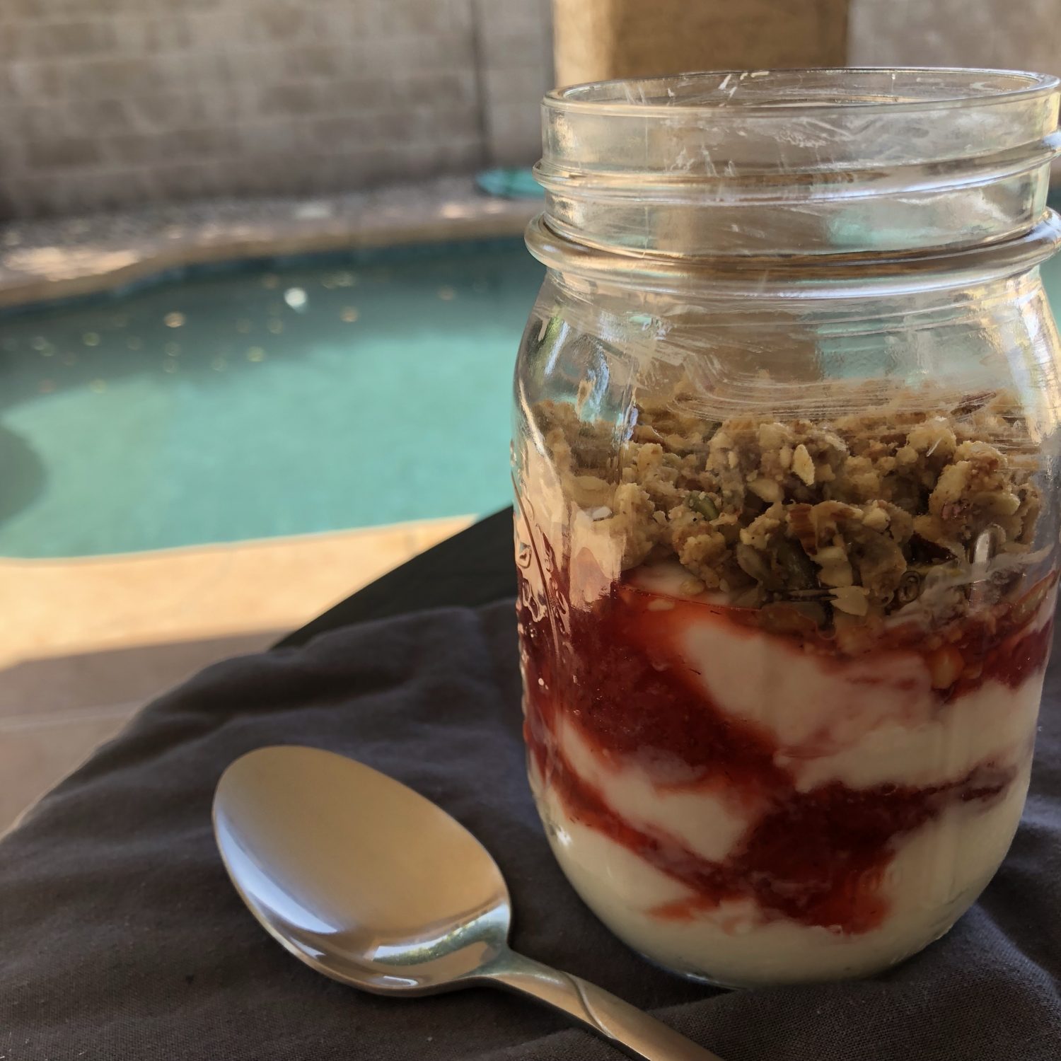 Granola parfait in a mason jar on a table with a pool in the background